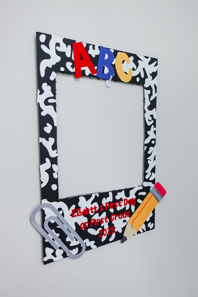 A black-and-white composition notebook-themed photo frame decorated with colorful cardstock cutouts, including red, blue, and yellow "ABC" letters, a glittery silver paperclip, and a layered paper pencil. The text reads โElliottโs First Day of First Grade 2025,โ making it a back-to-school photo prop.