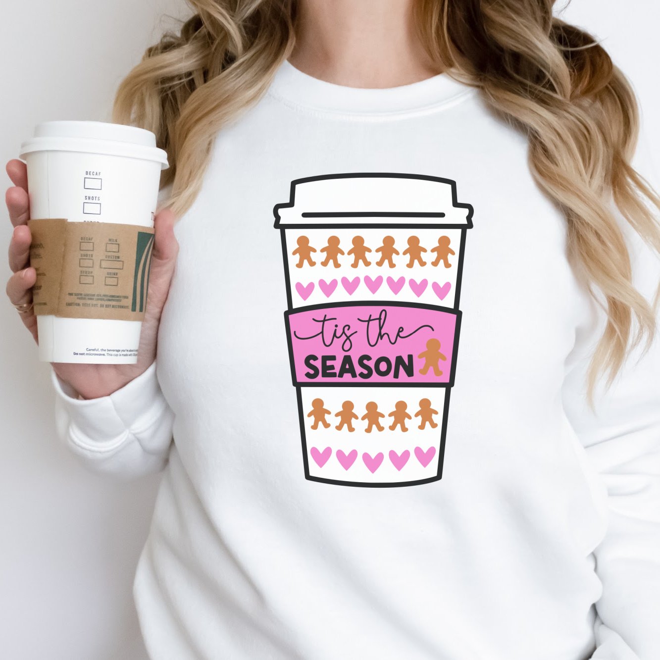 White sweatshirt with a pink and brown holiday coffee cup design featuring gingerbread men and hearts, held by a woman holding a matching coffee cup.