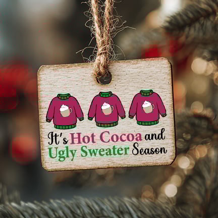 Wooden ornament with three illustrated sweaters and the phrase โItโs Hot Cocoa and Ugly Sweater Seasonโ hanging on a Christmas tree.