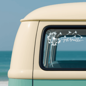 White vinyl decal of a dandelion blowing in the wind with the words “just breathe” on the window of a retro cream-and-teal van parked by the ocean.