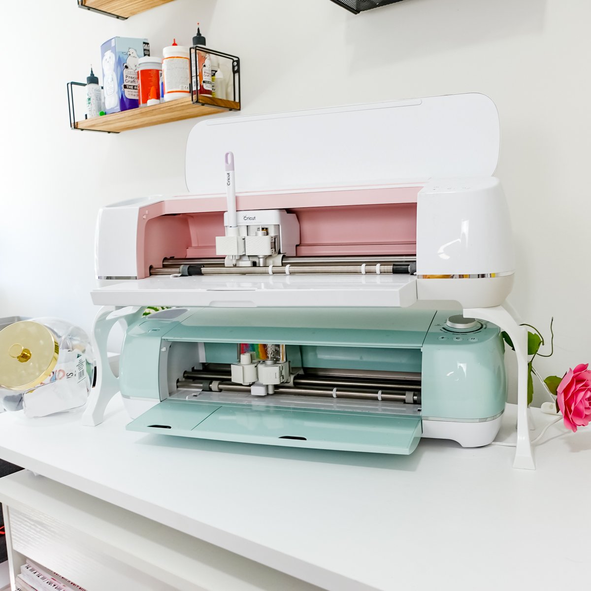 Cricut Maker and Cricut Explore Air 2 cutting machines displayed together in a bright craft room setup with supplies on shelves.